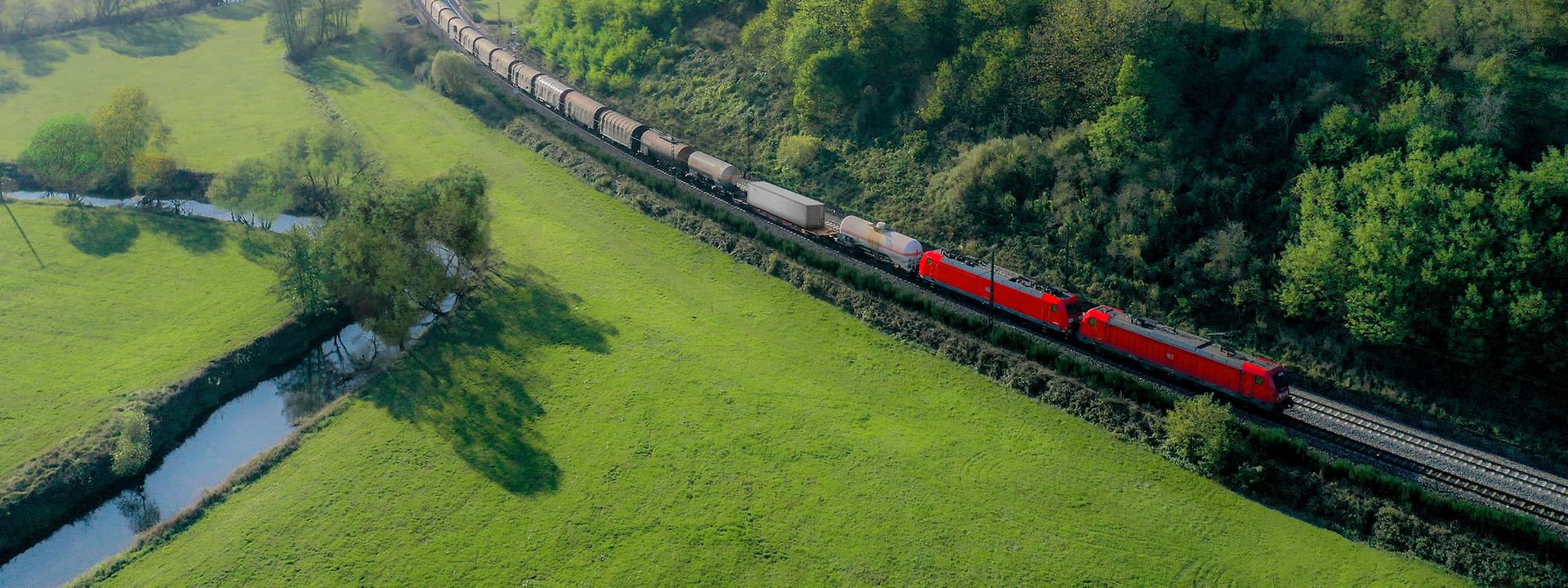 Red DB Cargo freight train travelling through a green hilly landscape with forests, meadows and a small stream